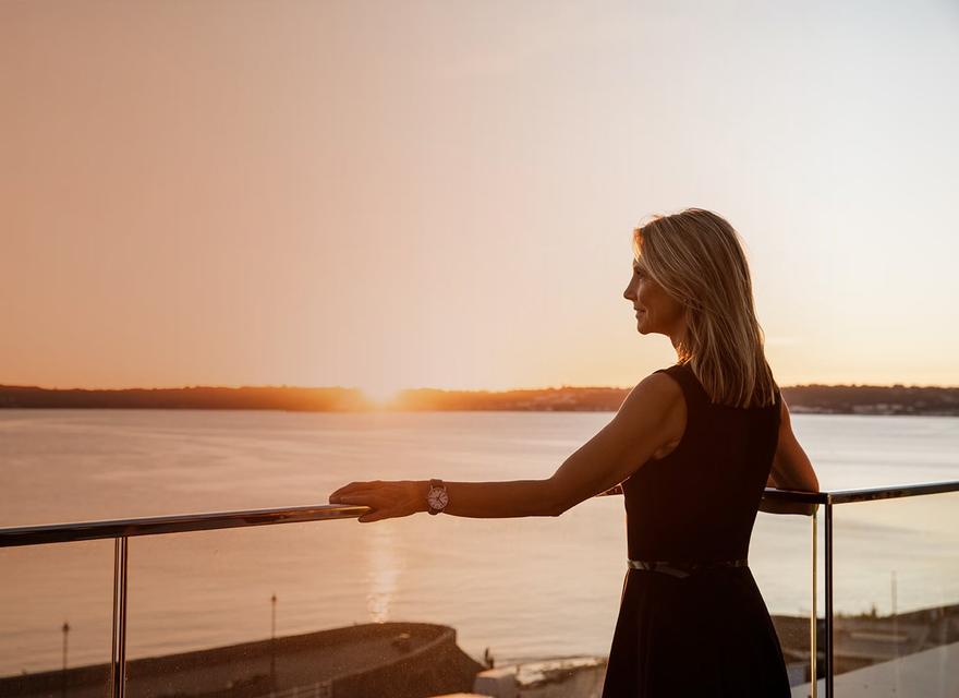 Businesswoman standing on office balcony overlooking St Aubin's bay at sunset