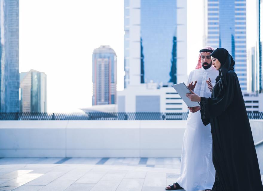 A man and woman in Arabic dress read a report together in front of high rise buildings