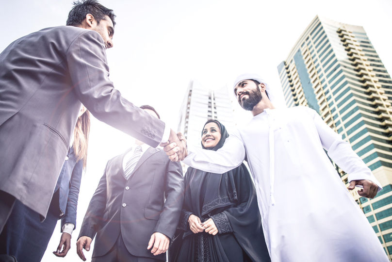 Two people shake hands, in front of other people and tall buildings.