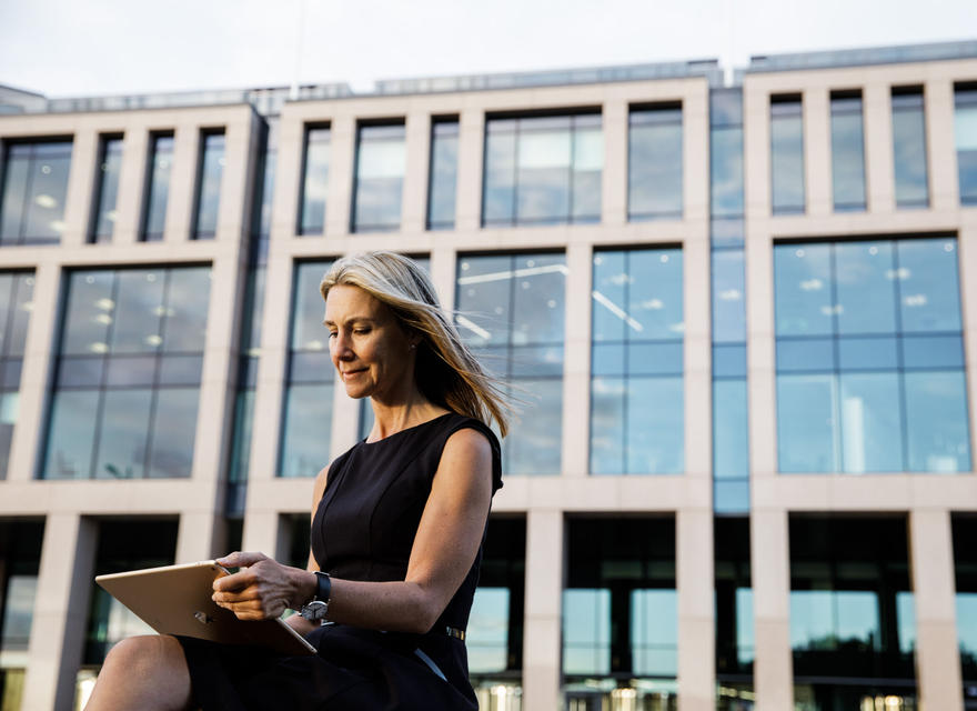 Woman works on a tablet outdoors in front of a corporate building.