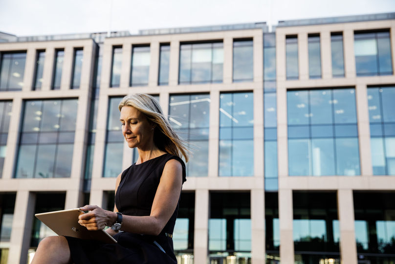 Woman works on a tablet outdoors in front of a corporate building.