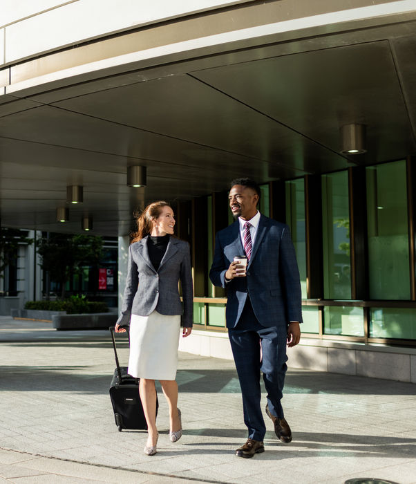 Two colleagues walking outdoors with coffee and luggage, deep in conversation.