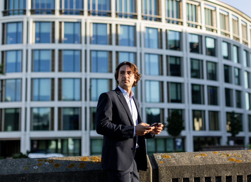 Man standing outside, looking up, holding a phone, with modern building in the background.