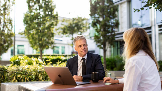 Man and woman seated outdoors at a business meeting, laptop and coffee cup in view.