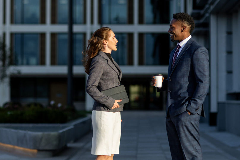 Two professionals talking while standing in a city plaza.