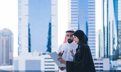 A man and woman in Arabic dress read a report together in front of high rise buildings