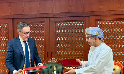 Two officials, one wearing a navy suit and the other traditional Omani dress, exchange red document folders during a formal signing ceremony, with Jersey and Oman flags displayed on the conference table.